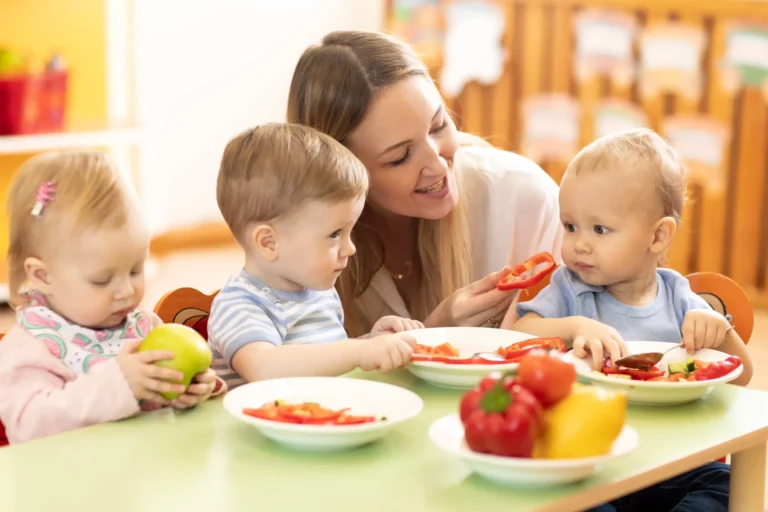 kindergarten teacher feeding toddlers with vegetables at table