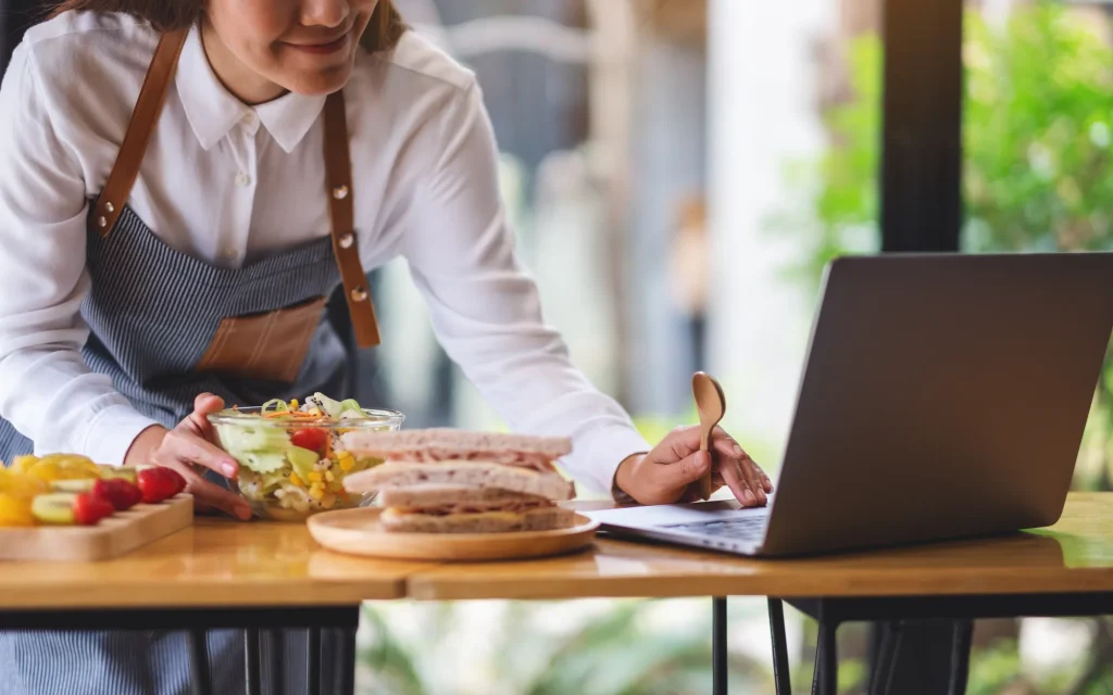 Frau mit Schürze bereitet Salat und Sandwiches auf einem Küchentisch zu und macht Notizen am Laptop